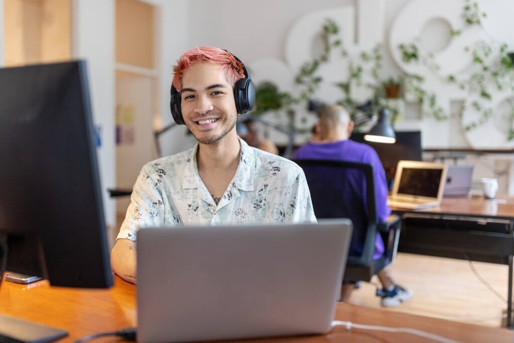 An online bachelor's degree student with dyed red hair and a nose piercing looks up from their laptop computer to smile and face the camera.